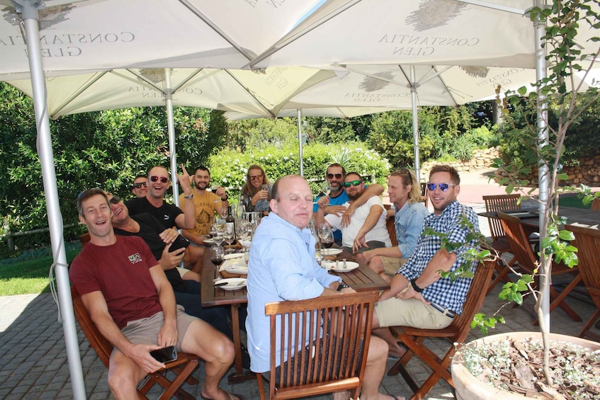 A group of people seated at an outdoor table under umbrellas, surrounded by greenery. 2