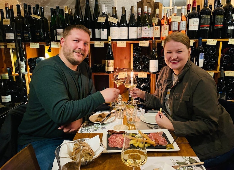 Deux personnes trinquent avec des verres à vin, assises à une table où se trouvent de la nourriture et des bouteilles de vin en arrière-plan. 1