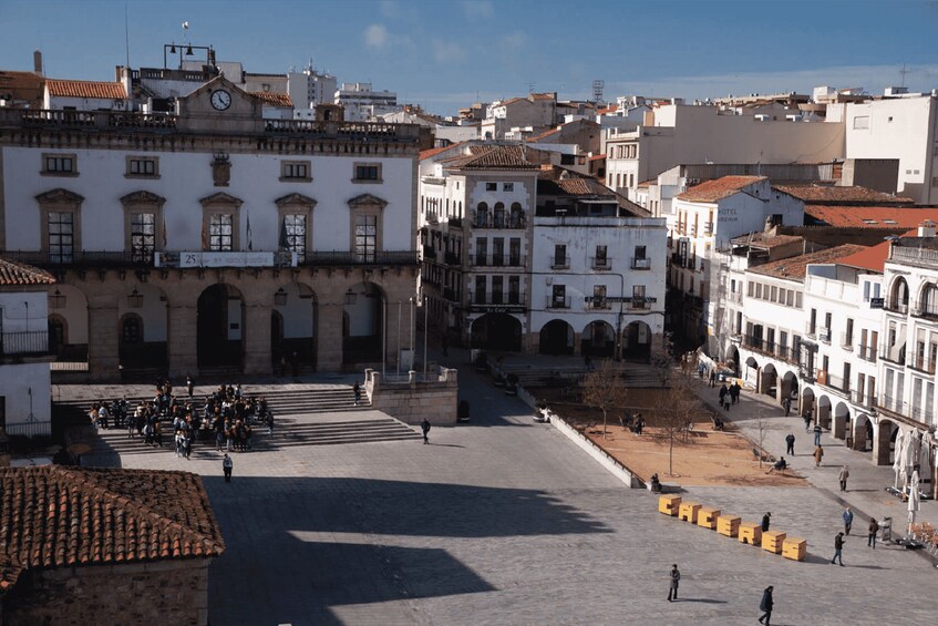 A historic square with buildings, a clock tower, and people walking. 1