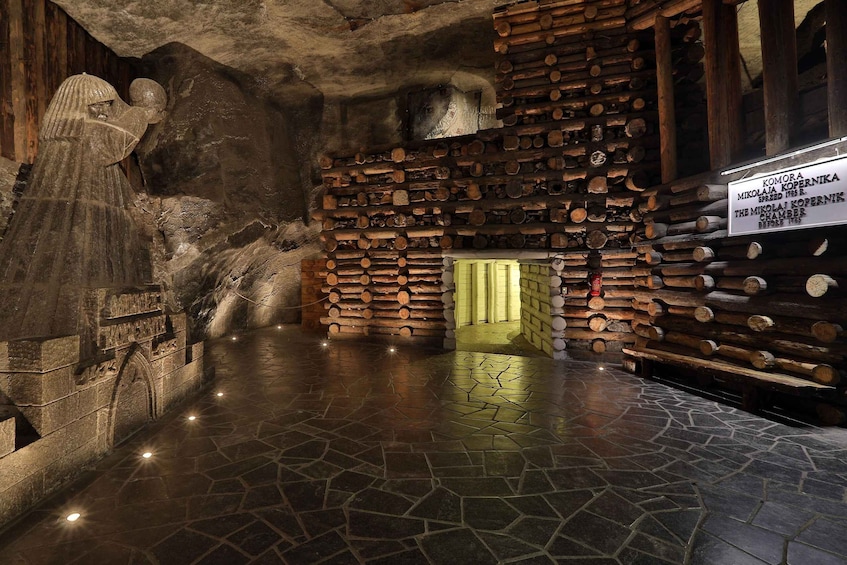 Habitación con paredes de piedra, un muro de troncos de madera, suelo de piedra y una puerta iluminada de verde. 1