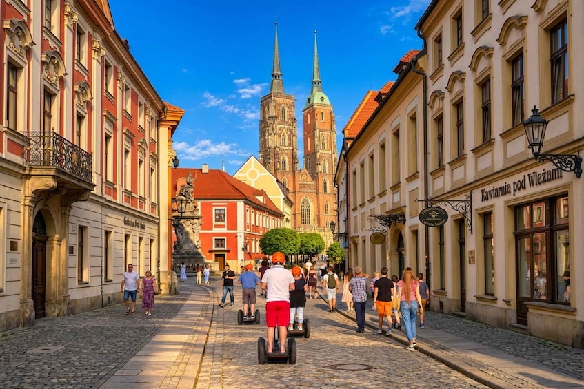 A cobblestone street lined with historic buildings, a church with a tall spire, and people walking and riding segways. 1