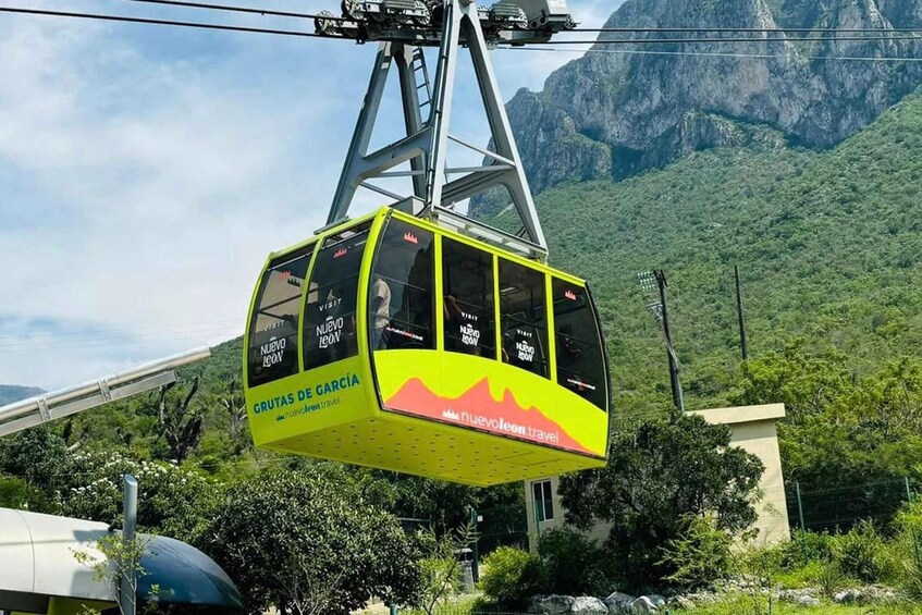 A yellow cable car with 'GRUTAS DE GARCÍA' and 'nuevo leon' on the side, suspended by cables against a mountainous backdrop. 1