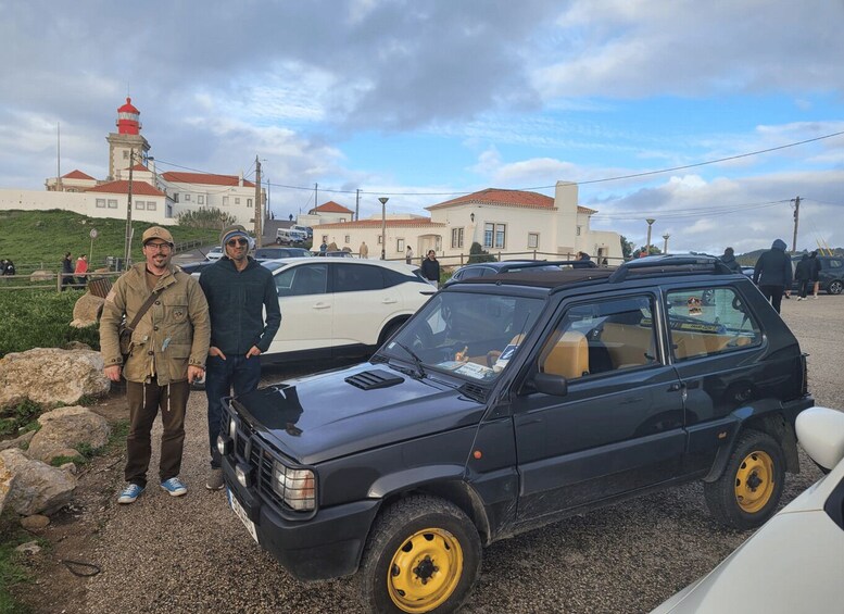 Two men standing beside a vintage car with yellow wheels, parked on a gravel surface with a lighthouse and buildings in the background. 1