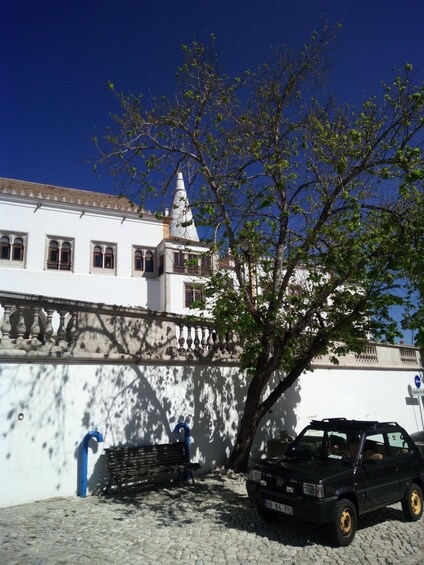 A white building with a tower, a tree, a bench, and a parked car. 5