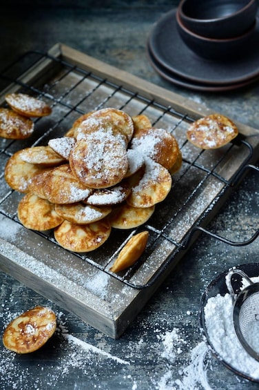 A tray of round, golden-brown pastries dusted with powdered sugar, resting on a wire rack. 1