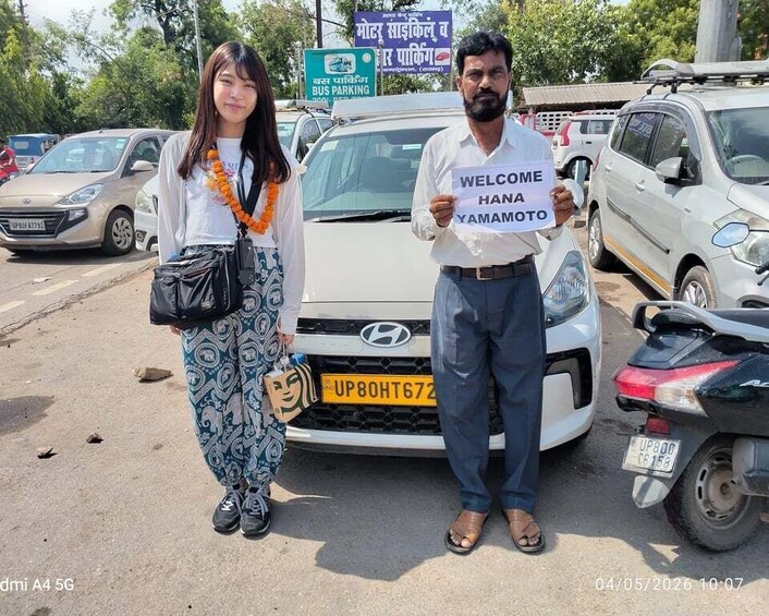 Two people standing in front of a white car with a sign that reads 'WELCOME HANA YAMAMOTO' and a bus parking sign in the background. 2