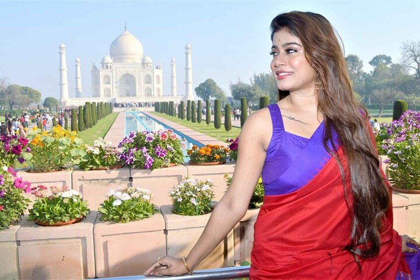 A person in a purple and red outfit standing near a garden with a view of the Taj Mahal. 2