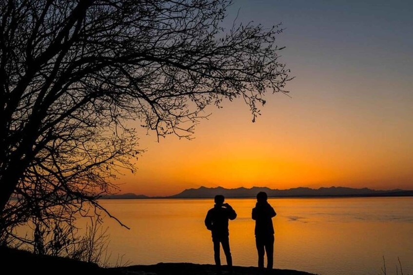 Kaksi ihmistä, joiden hahmot erottuvat selkeästi auringonlaskun taustaa vasten tyynen vesistön yllä, ja kauempana näkyy vuoria. 1