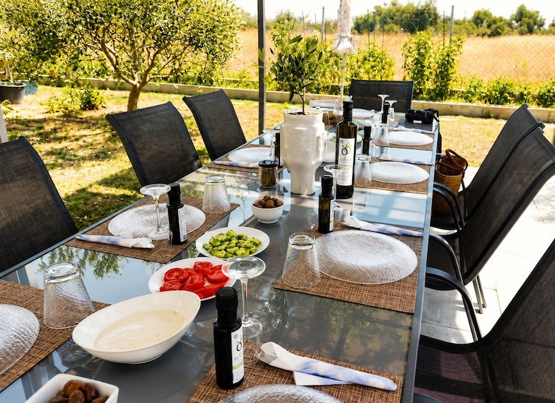 An outdoor dining table set with chairs, plates, bowls, and bottles, surrounded by greenery and trees. 2