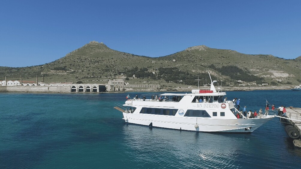 Een witte boot genaamd 'M.N. Intrepid' afgemeerd aan een pier met een bergachtige achtergrond. 1