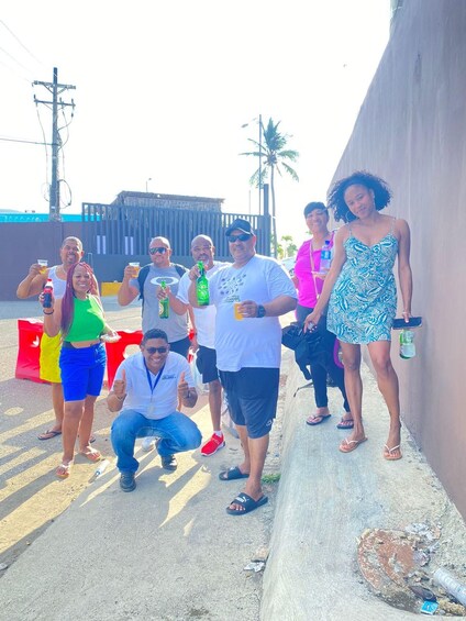A group of people standing outdoors, some holding drinks, with a fence and palm trees in the background. 2