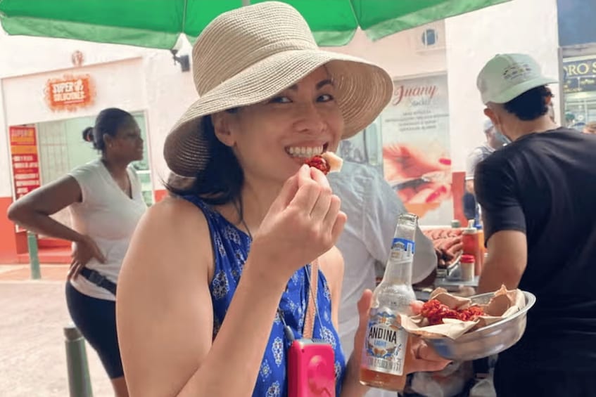 A person wearing a hat and a blue patterned top, holding a bottle of Andina liquor and a bowl of food. 2