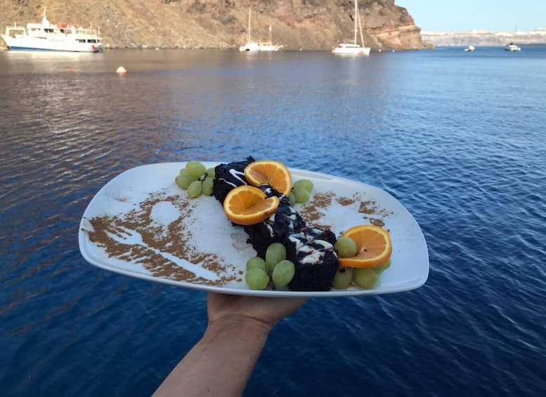 Un piatto di dessert con uva e fettine d'arancia su uno specchio d'acqua, sullo sfondo barche e scogliere. 5