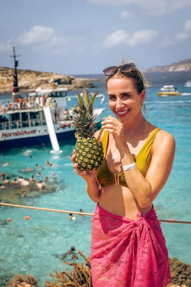 Henkilö, jolla on kädessään ananas, päällään keltainen bikinitoppi ja vaaleanpunainen hame, seisoo vesistön äärellä. Taustalla näkyy veneitä ja ihmisiä. 1