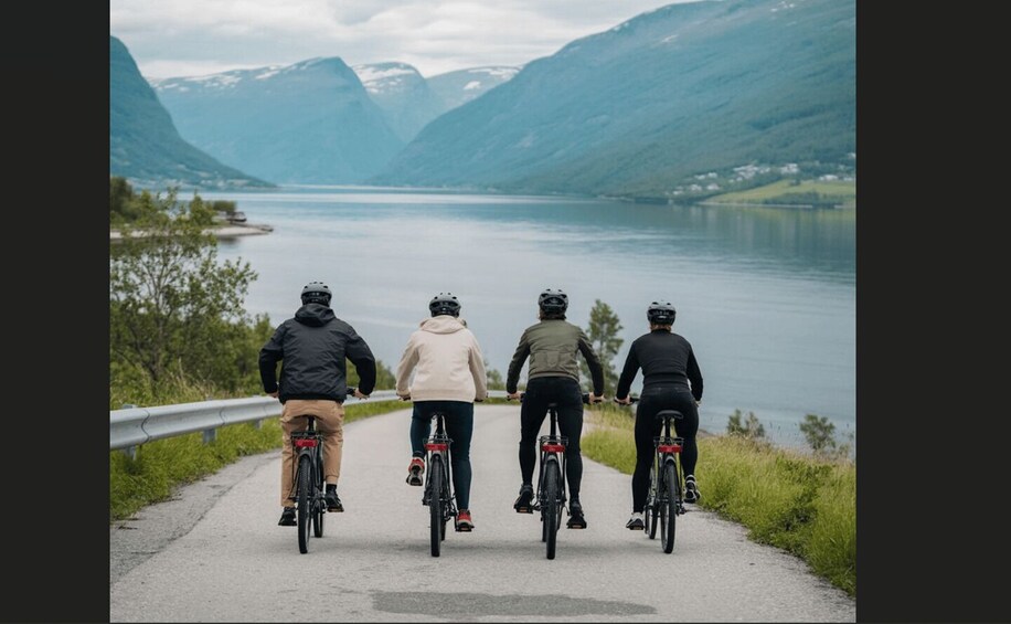 Four cyclists on a paved path beside a lake with mountains in the background. 3