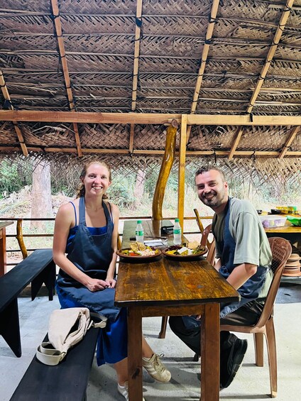 Two people sitting at a wooden table with food, under a thatched roof. 2