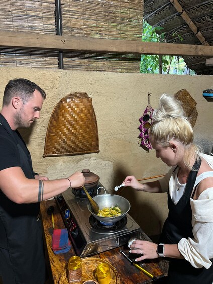Two people cooking in a rustic kitchen with a woven ceiling and a traditional stove. 5