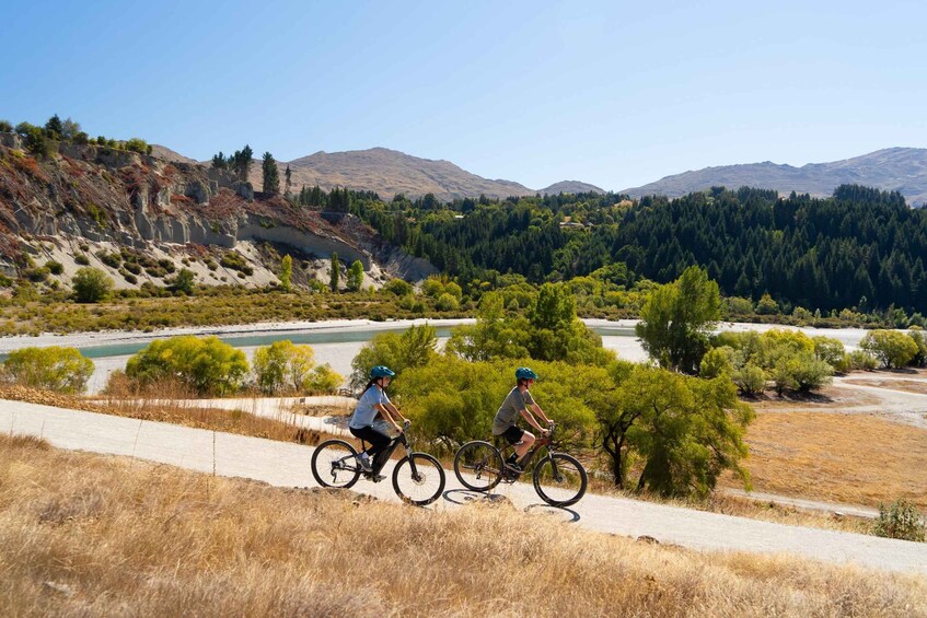 Two cyclists on a dirt path with a river and mountains in the background. 1