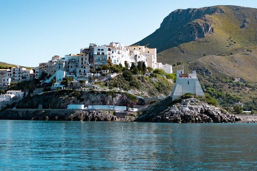 A coastal town with white buildings on a hillside, a rocky outcrop, and a mountain in the background. 4