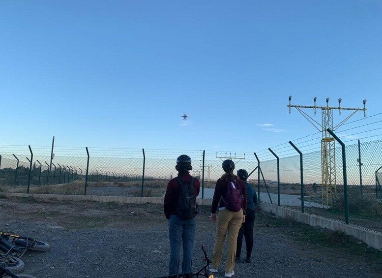 A group of people standing on a dirt path, looking at an airplane flying in the distance. 1