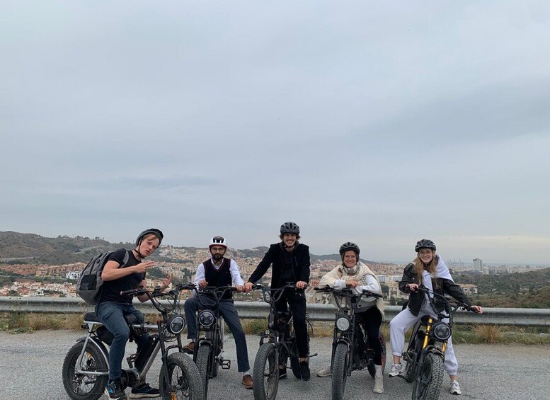 Five people on bicycles on a road with a cityscape in the background. 1