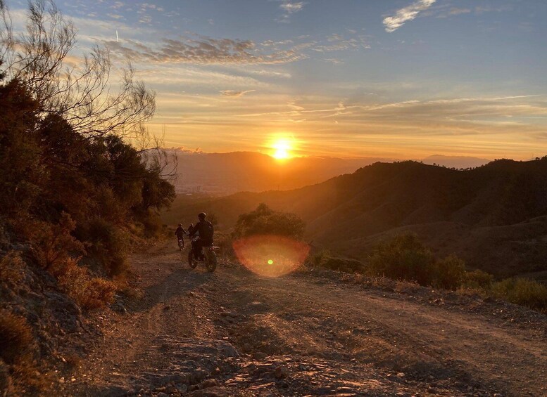 A dirt road with two motorcyclists riding away from the viewer, with a sunset in the background. 1