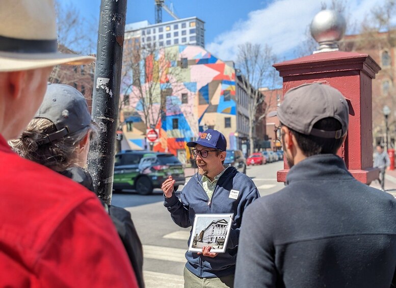 Une personne tenant une tablette sur laquelle est affichée une image d’un bâtiment, debout dans une rue où l’on aperçoit, en arrière-plan, des bâtiments aux couleurs vives. 2