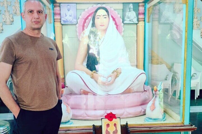 Ein Mann steht neben einer Statue einer sitzenden Figur in traditioneller Pose, mit einem rosafarbenen Kissen und einer kleinen Statue daneben. 3