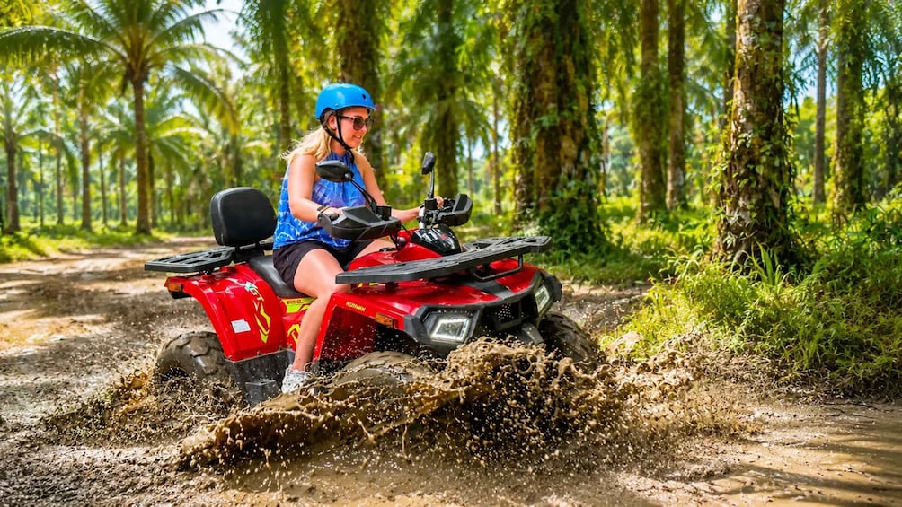 A person riding a red ATV on a muddy path surrounded by tropical trees. 1