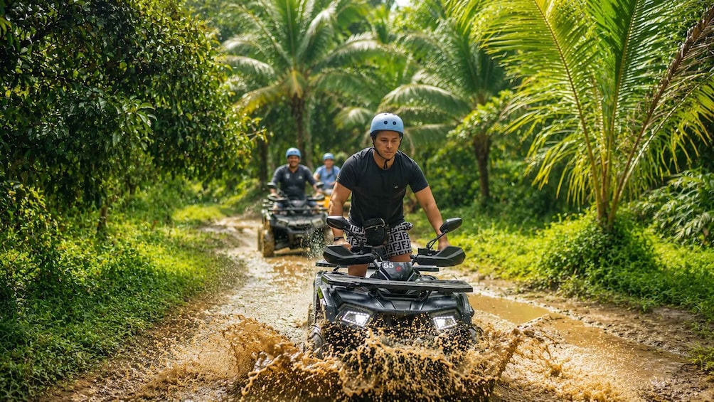 A group of people on all-terrain vehicles (ATVs) navigating a muddy path through a tropical forest. 3