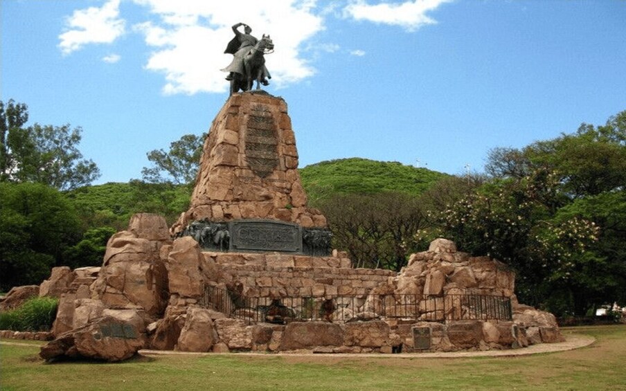 A stone monument with a statue of a person on horseback, surrounded by rocks and greenery. 5