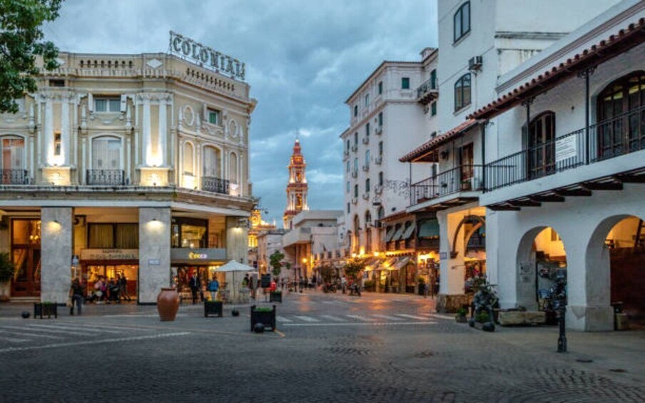 A cobblestone street lined with historic buildings, a prominent clock tower, and a mix of commercial and residential architecture. 1