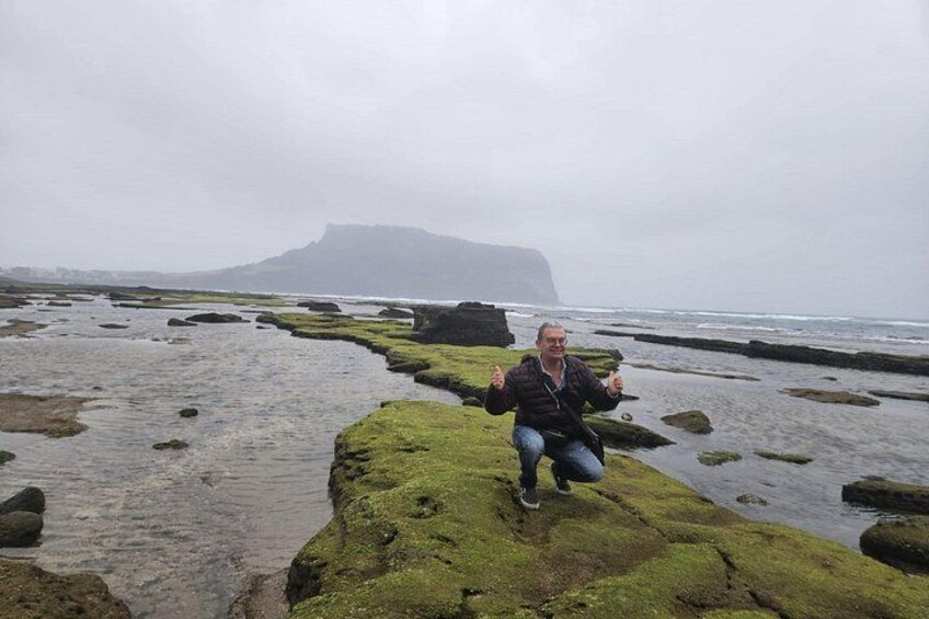 A person squatting on a moss-covered rock formation with a rugged coastline and a distant mountain in the background. 1