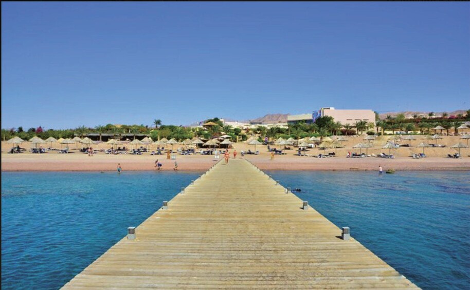 A wooden pier extending into calm waters, leading to a sandy beach with umbrellas and people. 2