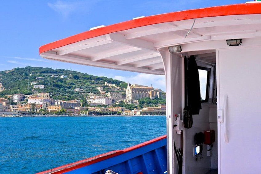 A boat with a red and white canopy, a blue railing, and a view of a coastal town with buildings and a hill in the background. 1