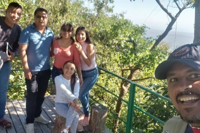 A group of people on a wooden deck with a green railing, overlooking a forested area with trees and a view of the ocean. 1