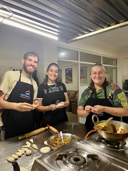 Three people in a kitchen preparing food, with a pot on the stove and dumplings on the counter. 2