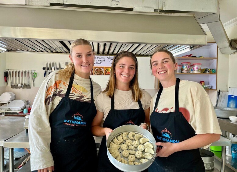 Three people in a kitchen holding a pot of dumplings, wearing aprons with 'Kathmandu Cooking Academy' text. 1