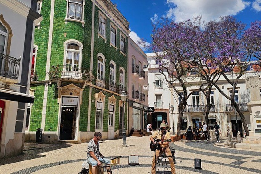 Une scène de rue avec des bâtiments, un arbre aux fleurs violettes et une personne qui joue de la guitare. 4