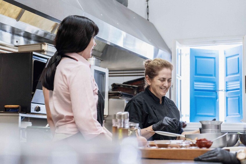 Una cocina con electrodomésticos de acero inoxidable, una persona con delantal de chef negro preparando alimentos, y otra persona con camisa rosa observando. 5