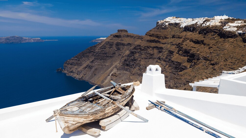 A white building with a wooden boat and a spear on the roof, overlooking a blue sea and a rocky island with white buildings. 2
