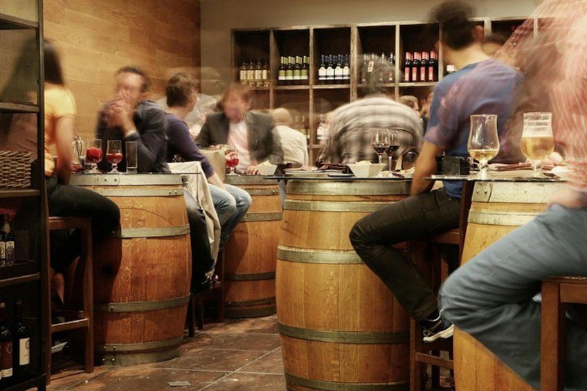 Eine Bar mit Holzfässern, Personen, die an Tischen sitzen, und Flaschenregalen. 1