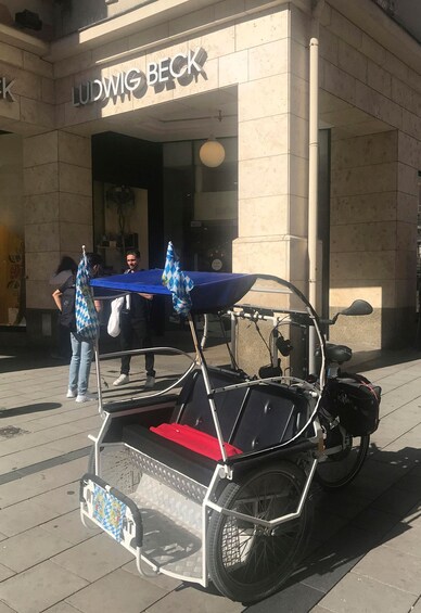 A custom-built motorcycle with a blue canopy and a red seat parked in front of a building with a sign that reads 'LUDWIG BECK'. 2