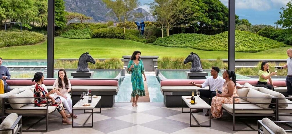 Eine Gruppe von Menschen sitzt auf Gartensofas und -stühlen, während eine Frau in einem grünen Kleid zwischen zwei Pferdestatuen geht. 1