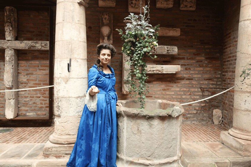 A person in a blue dress standing in front of a stone fountain with a hanging plant. 5
