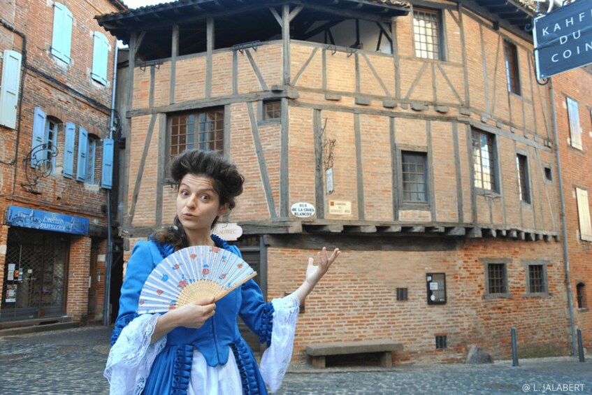 A person in historical costume holding a fan in front of a half-timbered building. 4