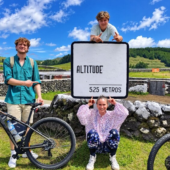 Zwei Personen halten ein Schild, auf dem 'ALTITUDE 525 METROS' steht, mit einem Fahrrad und einer Steinmauer im Hintergrund. 4