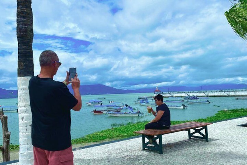 A man taking a photo of another man sitting on a bench by the water with boats in the background. 4