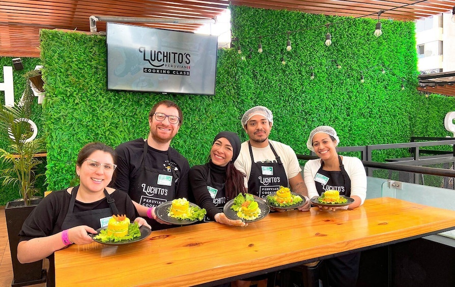 Cinque persone con grembiuli neri, con il logo 'Luchito's Peruvian Cooking Class', mostrano piatti preparati. 4