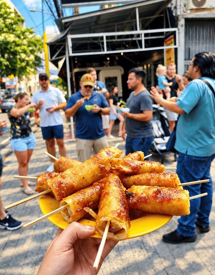 A person holding a plate of skewered food, likely a street food snack, in an outdoor setting with people around. 1